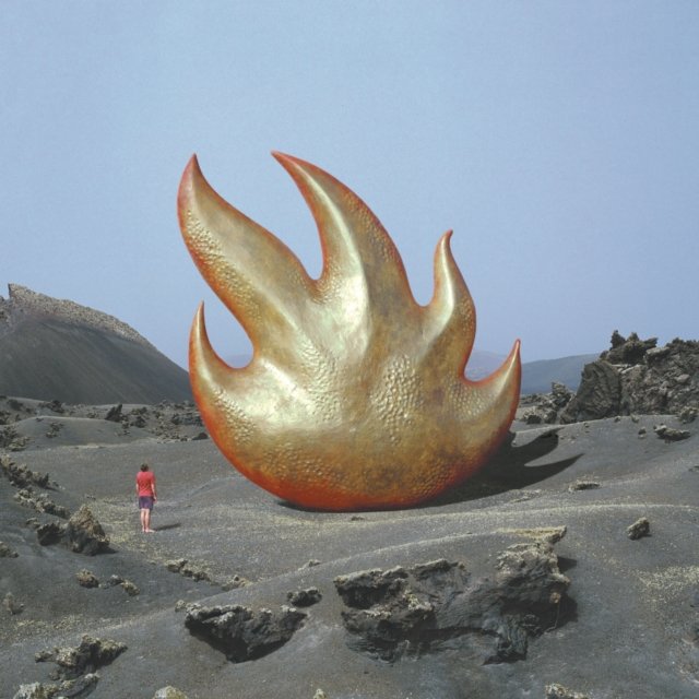 A giant golden flame sculpture towering over a person in a volcanic ash landscape.