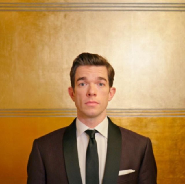 Comedian John Mulaney wearing a dark suit against a textured gold background.
