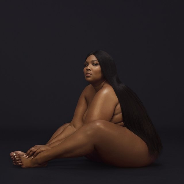 Artistic nude studio portrait of singer Lizzo seated against a black background.