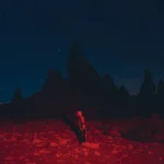 A skeleton standing in a red glowing desert under a dark, starry night sky.