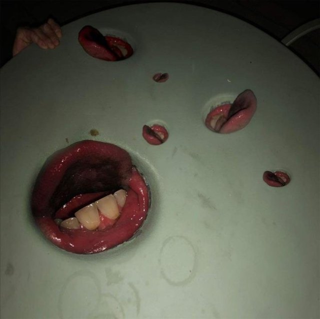 Surreal photo of human mouths and tongues protruding through holes in a plastic table.