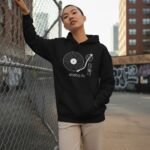 Woman with a buzz cut wearing a black analog life hoodie by a fence.