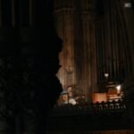A musician plays acoustic guitar in a dimly lit Gothic cathedral near a pipe organ.