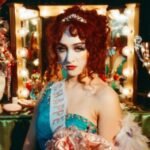 Chappell Roan in pageant attire holding roses in front of an illuminated vanity mirror.