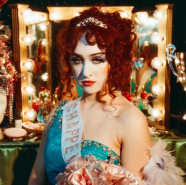 Chappell Roan in pageant attire holding roses in front of an illuminated vanity mirror.