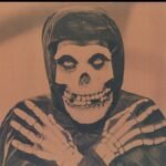 Sepia portrait of the Crimson Ghost wearing a skull mask and skeletal gloves.