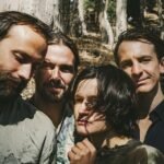 Close-up group portrait of four people standing in a sun-dappled forest.