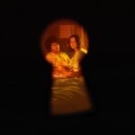 Keyhole view of a couple sitting on a bed in a warmly lit room.