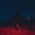 Person in a skeleton suit standing on a red-lit desert facing dark mountains.