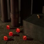 Five translucent red dice scattered on dark asphalt in a moody alleyway.
