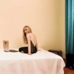 Sabrina Carpenter sitting on a bed looking over her shoulder in a backless dress.